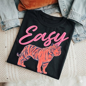 May include: Black t-shirt with the word "Easy" in pink script above a coral and navy tiger illustration. The shirt is displayed on a cream-colored surface with a denim jacket and a brown pillow in the background.