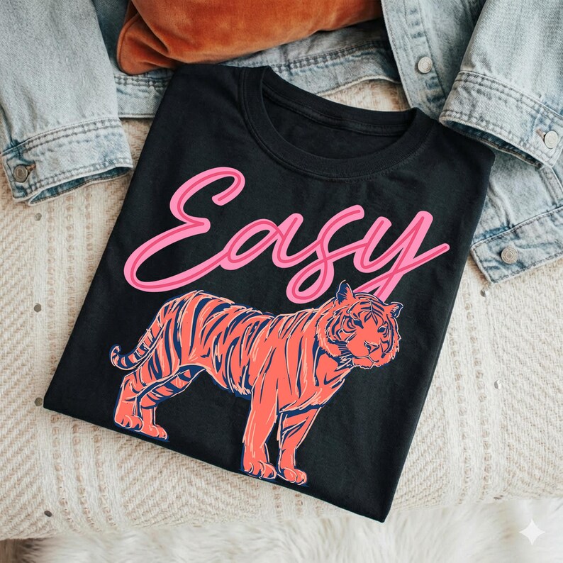 May include: Black t-shirt with the word "Easy" in pink script above a coral and navy tiger illustration. The shirt is displayed on a cream-colored surface with a denim jacket and a brown pillow in the background.