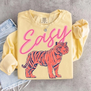 May include: A light yellow sweatshirt with the word "Easy" in pink script above a coral and navy tiger illustration. The sweatshirt has long sleeves and a crew neck. A silver necklace is visible.