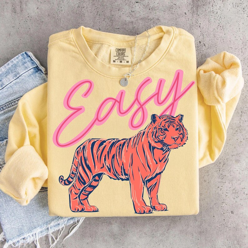 May include: A light yellow sweatshirt with the word "Easy" in pink script above a coral and navy tiger illustration. The sweatshirt has long sleeves and a crew neck. A silver necklace is visible.