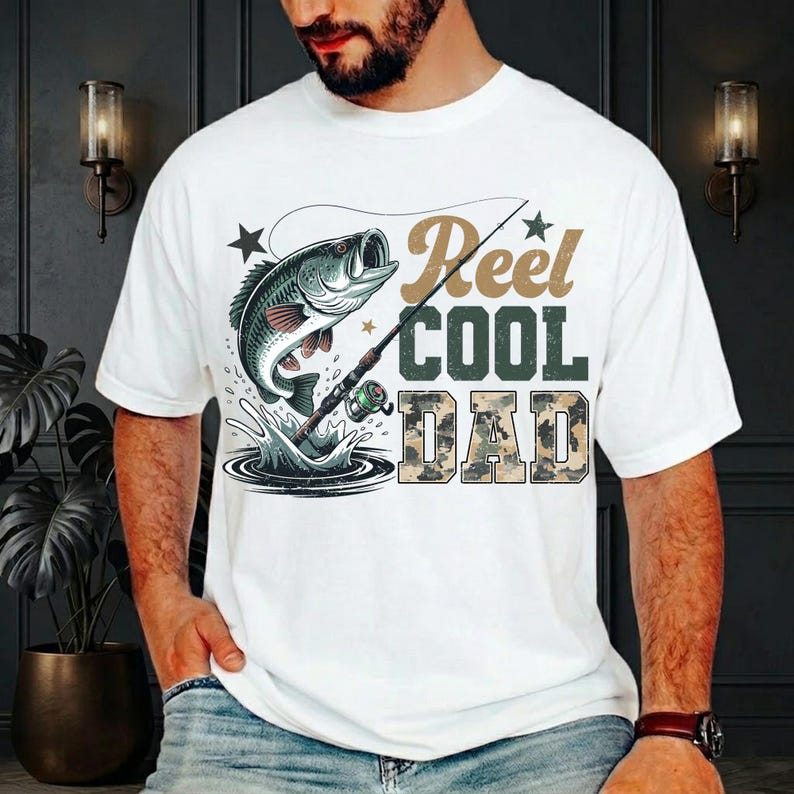 Może przedstawiać: Biała koszulka z grafiką przedstawiającą rybę wyskakującą z wody z wędką. Tekst "Reel COOL DAD" jest wydrukowany w kolorach brązowym, zielonym i moro. Świetny prezent dla wędkarzy.