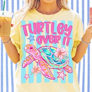 May include: Yellow t-shirt with the text "TURTLEY OVER IT" in pink. The shirt features a pink and teal turtle graphic with hibiscus flowers and stars. The shirt has light blue stripes.