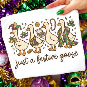 Op de afbeelding: Wit T-shirt met een cartoon illustratie van vier ganzen in Mardi Gras kostuums. De ganzen dragen maskers, kralen en een gevederde boa. De tekst "just a festive goose" is geschreven in een scriptlettertype.