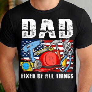 May include: Black t-shirt with a graphic featuring the word "DAD" in white block letters above an American flag design. The flag design includes various tools and the text "FIXER OF ALL THINGS".
