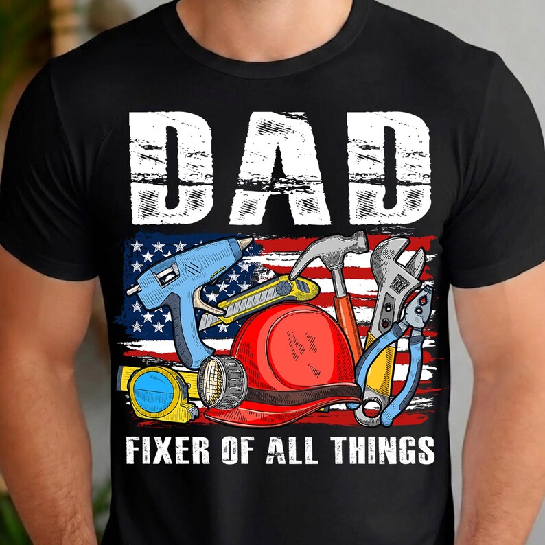 May include: Black t-shirt with a graphic featuring the word "DAD" in white block letters above an American flag design. The flag design includes various tools and the text "FIXER OF ALL THINGS".