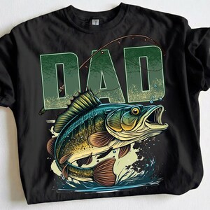 Pu&ograve; includere: T-shirt nera con una grafica di un pesce che salta fuori dall'acqua e la parola "DAD" in verde sopra. Il pesce &egrave; dettagliato con squame blu, verdi e marroni, e una canna da pesca &egrave; arcuata sopra.