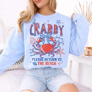 May include: Light blue sweatshirt with a crab graphic. The graphic includes a red crab with a blue bow, seashells, and the text "If Crabby Feeling Beachy Please Return to the Beach."