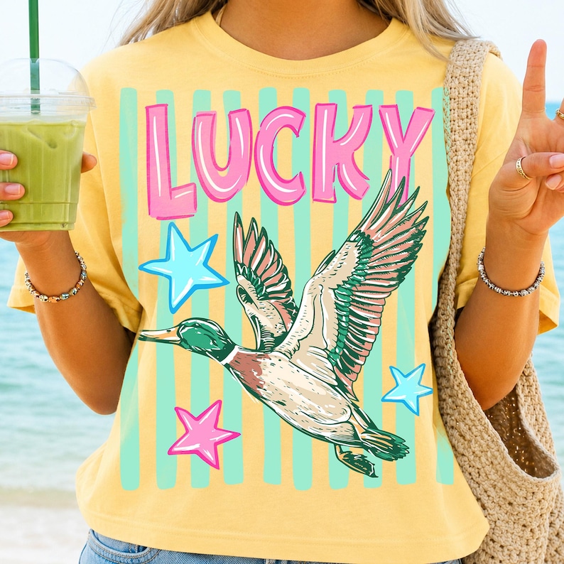 Puede incluir: Camiseta amarilla con la palabra "LUCKY" en letras rosas con contorno azul, un dibujo de un pato volando y estrellas azules y rosas. La camiseta tiene un corte holgado y mangas cortas.