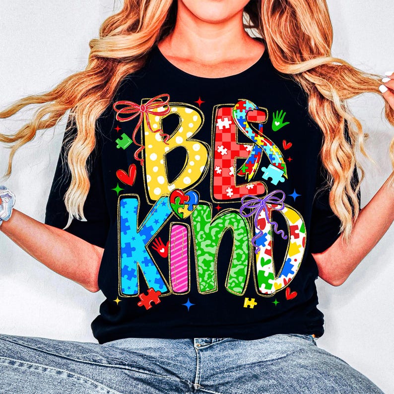 May include: Black t-shirt with the words "BE KIND" in colorful, puzzle-piece-themed letters. The design includes hearts, stars, and puzzle pieces, promoting autism awareness. The shirt is a crew neck.