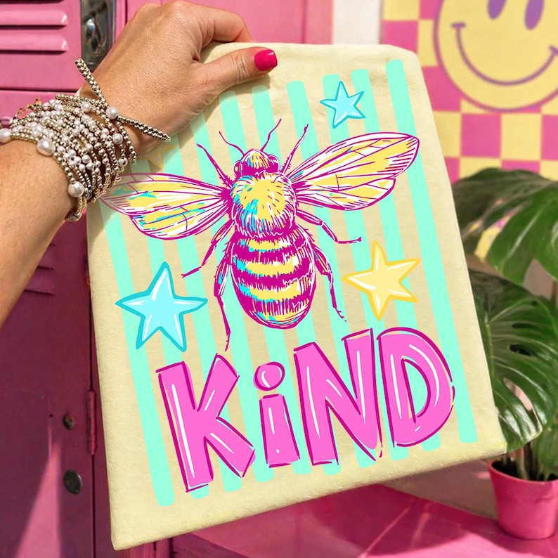 Puede incluir: Una bolsa de mano de color amarillo claro con una gran ilustraci&oacute;n de abeja colorida, rayas turquesas y la palabra "KIND" en rosa. La abeja tiene alas amarillas y rosas y un cuerpo a rayas. Tambi&eacute;n se presentan estrellas azules y amarillas.