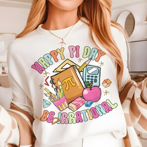 May include: White sweatshirt with a colorful graphic design that says "Happy Pi Day Be Irrational". The graphic includes a notebook with a pi symbol, pencils, a calculator, a protractor, a ruler, and an apple.