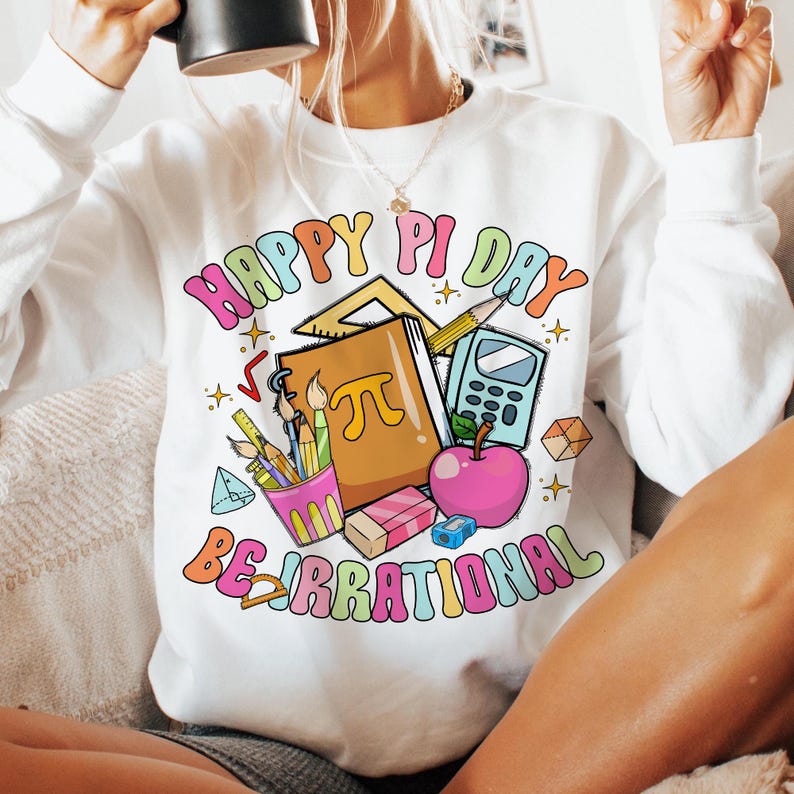 May include: White sweatshirt with a colorful graphic design that says "Happy Pi Day Be Irrational". The graphic includes a notebook, a calculator, pencils, a protractor, and an apple.