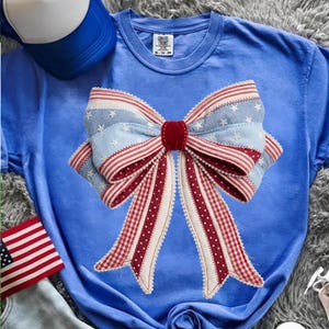 May include: A blue t-shirt featuring a large bow design. The bow has red and white striped ribbons, light blue sections with white stars, and a red velvet center. The shirt is a solid blue color.