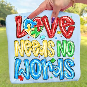 May include: Light blue t-shirt with the words "LOVE NEEDS NO WORDS" in colorful, outlined letters. The word "LOVE" features a heart-shaped puzzle piece design. The shirt is held up by a hand.