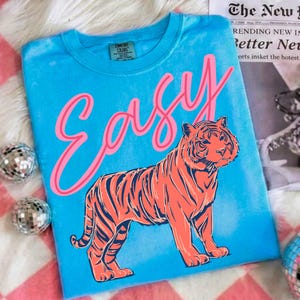 May include: A turquoise t-shirt with the word "EASY" in pink script and a large orange tiger illustration. The shirt has a crew neck and a Comfort Colors tag. The background includes disco balls and a newspaper.