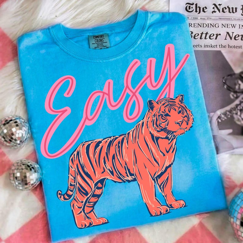 May include: A turquoise t-shirt with the word "EASY" in pink script and a large orange tiger illustration. The shirt has a crew neck and a Comfort Colors tag. The background includes disco balls and a newspaper.