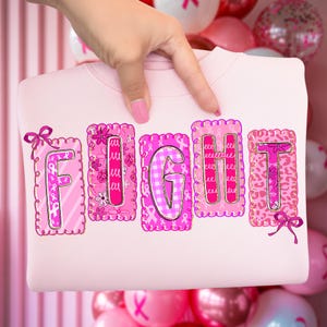 May include: A pale pink sweatshirt featuring the word "FIGHT" in pink, adorned with ribbons and floral designs. The letters are framed with decorative borders. The sweatshirt is held up by a hand.