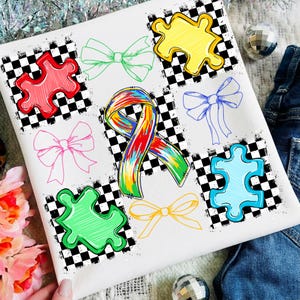 May include: White t-shirt with a design of colourful puzzle pieces and bows. The design includes a rainbow ribbon and various puzzle pieces in red, yellow, green, and blue, along with several bow illustrations. The background has a black and white checkered pattern.