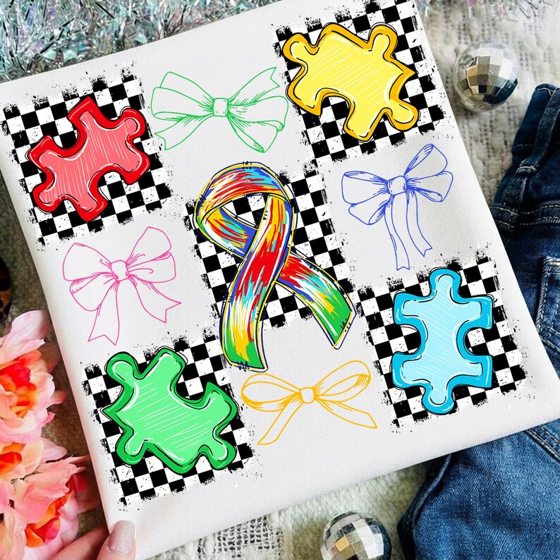 May include: White t-shirt with a design of colourful puzzle pieces and bows. The design includes a rainbow ribbon and various puzzle pieces in red, yellow, green, and blue, along with several bow illustrations. The background has a black and white checkered pattern.