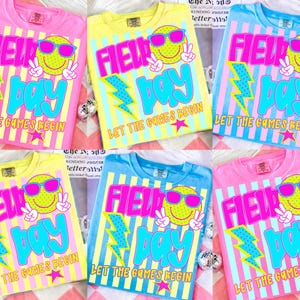 Puede incluir: Varias camisetas coloridas con un diseño de "Field Day". Cada camiseta presenta una carita sonriente con gafas de sol, un rayo y el texto "Let the Games Begin". Las camisetas son rosas, amarillas y azules.