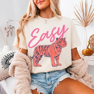 May include: Cream-colored t-shirt with the word "Easy" in pink script above a coral-colored tiger illustration with navy blue stripes. The shirt is worn with denim shorts.