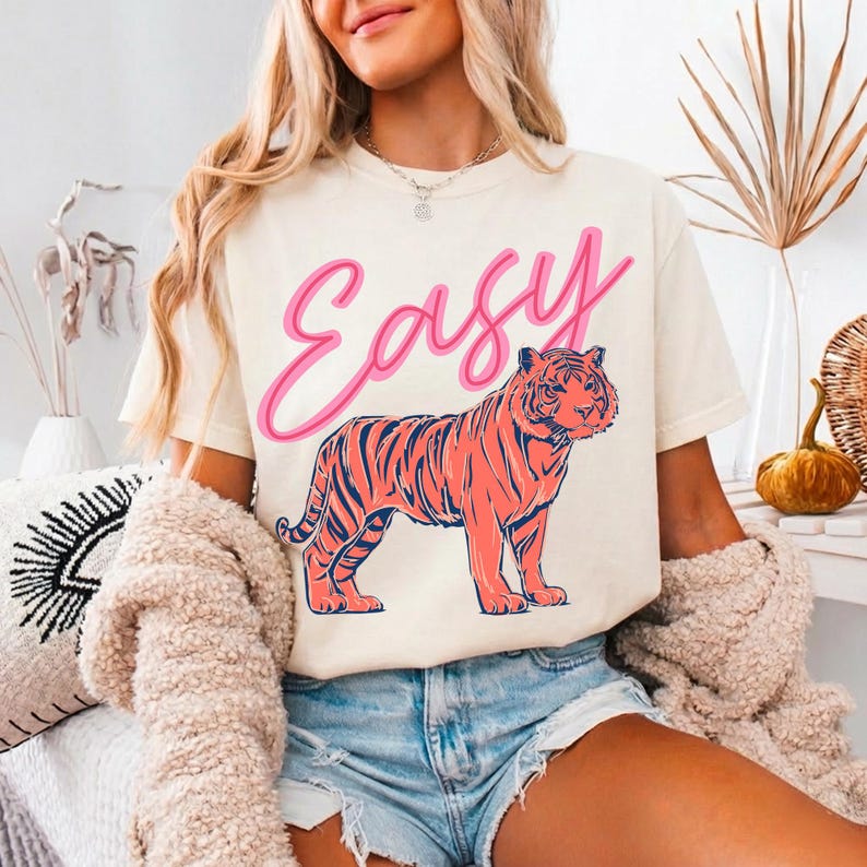 May include: Cream-colored t-shirt with the word "Easy" in pink script above a coral-colored tiger illustration with navy blue stripes. The shirt is worn with denim shorts.