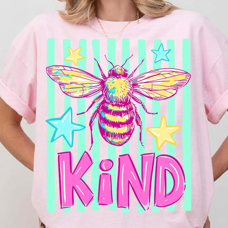 Puede incluir: Camiseta rosa claro con un dise&ntilde;o de abeja colorida, estrellas y la palabra "KIND" en rosa. La abeja tiene alas amarillas y azules, y la camiseta tiene un fondo a rayas verde claro. Dise&ntilde;o alegre.