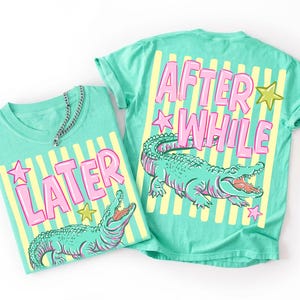 Può includere: Due magliette turchesi con un design grafico. Una dice "LATER" e l'altra "AFTER WHILE" in lettere rosa. Entrambe presentano un'illustrazione di un alligatore verde e strisce gialle e bianche.