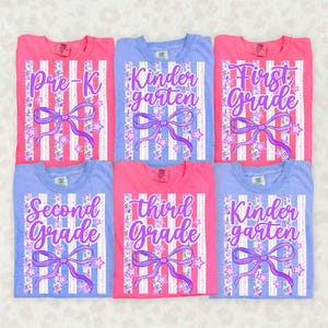Op de afbeelding: Zes kleurrijke t-shirts met tekst over schoolniveau. De shirts zijn roze en blauw, met witte verticale strepen, paarse strikken en steraccenten. De tekst bevat "Pre-K", "Kindergarten", "First Grade", "Second Grade" en "Third Grade".