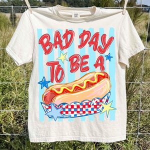 以下が含まれることがあります： 「BAD DAY TO BE A」の赤い文字とホットドッグのイラストが描かれた、クリーム色のTシャツ。水色のストライプと星のアクセントがデザインを完成させています。Tシャツは物干しロープに吊るされています。