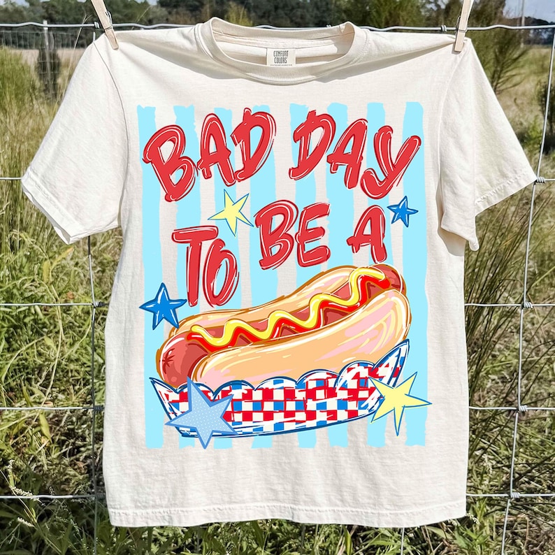 以下が含まれることがあります： 「BAD DAY TO BE A」の赤い文字とホットドッグのイラストが描かれた、クリーム色のTシャツ。水色のストライプと星のアクセントがデザインを完成させています。Tシャツは物干しロープに吊るされています。