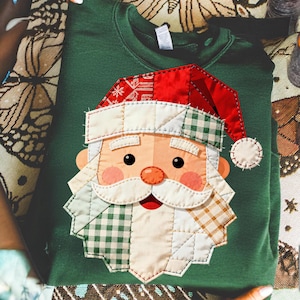 Peut inclure: Un sweat-shirt vert foncé avec un motif de Père Noël en patchwork. Le visage du Père Noël comprend un bonnet rouge avec des détails de flocons de neige blancs, une barbe blanche et des joues roses. Le patchwork utilise des motifs à carreaux et unis.