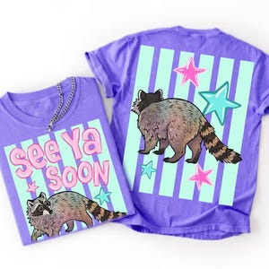 May include: Two purple t-shirts with raccoon graphics. One shirt has the text "See Ya Soon" with a raccoon illustration. The other shirt features a raccoon, stars, and vertical stripes. The shirts are casual wear.