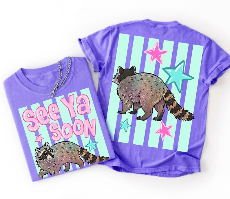 May include: Two purple t-shirts with raccoon graphics. One shirt has the text "See Ya Soon" with a raccoon illustration. The other shirt features a raccoon, stars, and vertical stripes. The shirts are casual wear.
