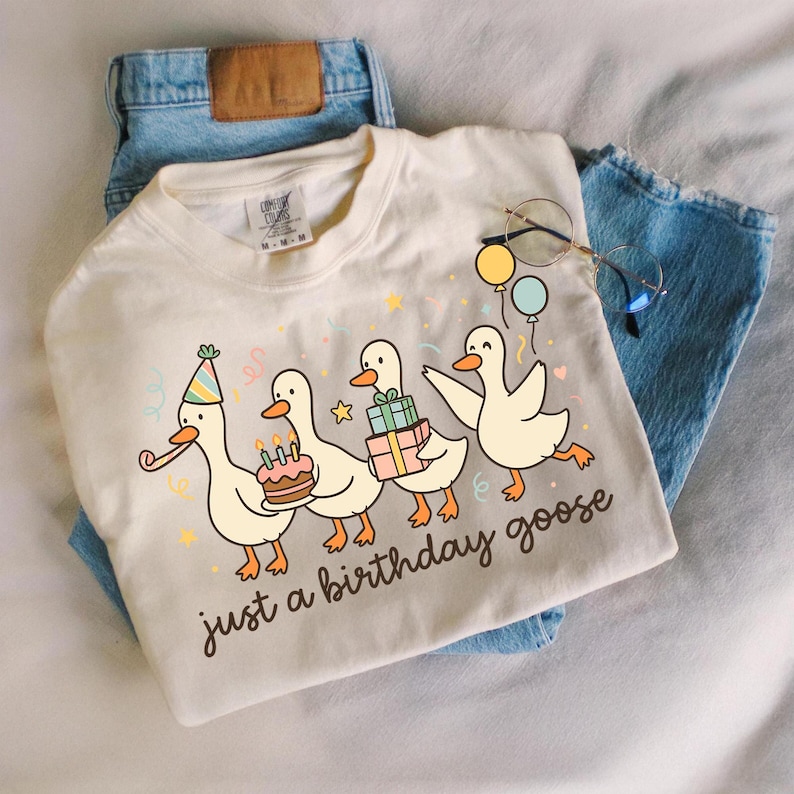 May include: Cream-colored t-shirt with a whimsical illustration of four cartoon geese celebrating a birthday. The geese are carrying a cake, gifts, and balloons. The text "just a birthday goose" is printed below the geese. The shirt is paired with blue jeans.