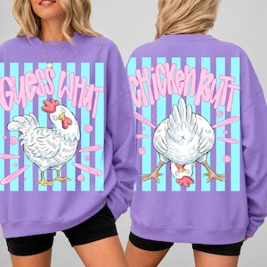 May include: A purple sweatshirt with a cartoon chicken design. The front of the sweatshirt says "Guess What" with a drawing of a chicken. The back says "Chicken Butt" with a chicken graphic. The background has blue and white stripes.