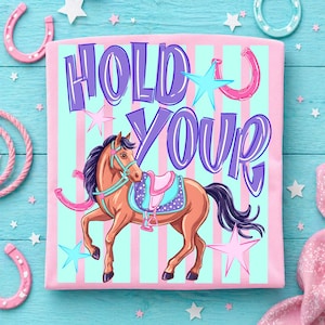 Pode incluir: Camiseta rosa com uma ilustração de cavalo de desenho animado e o texto "HOLD YOUR". O cavalo é marrom com crina e cauda pretas, usando uma sela. O fundo tem listras rosa e azuis, com estrelas e ferraduras.