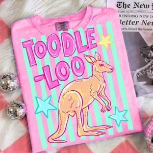 May include: Pink t-shirt with the words "TOODLE LOO" in pink, a kangaroo illustration, and star graphics. The shirt has vertical stripes in light blue and is made of Comfort Colors material.