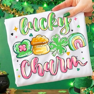 Puede incluir: Camiseta blanca con las palabras "Lucky Charm" en letras verdes y rosas. La camiseta presenta ilustraciones coloridas de un corazón verde, una olla de oro, un trébol de cuatro hojas y un arcoíris.