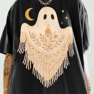 May include: Dark gray t-shirt featuring a cream-colored ghost design with a macrame shawl. The ghost has two dark brown eyes and is adorned with a gold crescent moon and three gold stars. The shawl has a floral pattern and fringe.