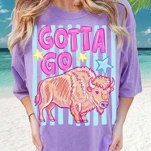 Può includere: Maglietta lilla con la scritta rosa "GOTTA GO", un'illustrazione di bisonte dei cartoni animati, stelle gialle e una stella blu. Il design è posto su uno sfondo a righe.