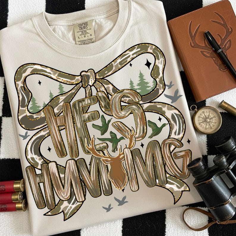 May include: Cream-colored t-shirt with the text "HE'S HUNTING" in a camouflage design, featuring a deer head and bow. The shirt has a bow design with green trees and birds. The shirt is on a black and white checkered surface.