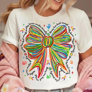 May include: White t-shirt featuring a colorful, hand-drawn bow design. The bow is composed of various colors and surrounded by words like "brave," "creative," and "kind." The design includes handprints, ribbons, and crosses.