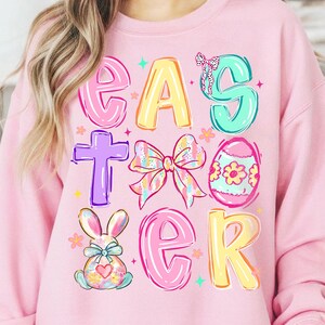 May include: Pink sweatshirt with the word "EASTER" in colorful, playful letters. The design includes a cross, a bow, an Easter egg, and a bunny, all in pastel colors. The sweatshirt is a comfortable, casual style.