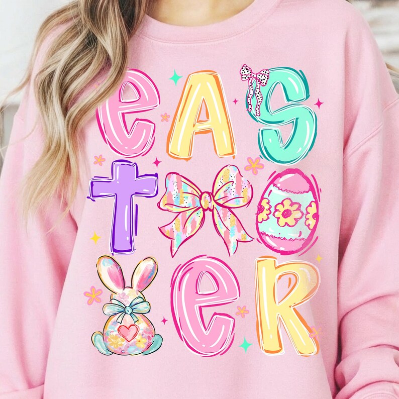 May include: Pink sweatshirt with the word "EASTER" in colorful, playful letters. The design includes a cross, a bow, an Easter egg, and a bunny, all in pastel colors. The sweatshirt is a comfortable, casual style.
