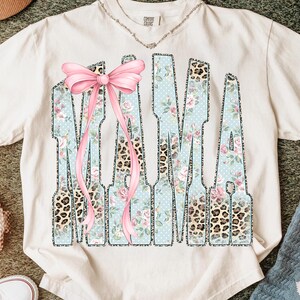 May include: Cream-colored t-shirt with the word "MAMA" in a unique design. The letters feature a light blue background with white polka dots and floral patterns, accented with leopard print and a pink bow.