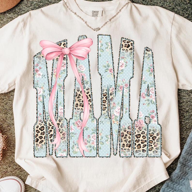May include: Cream-colored t-shirt with the word "MAMA" in a unique design. The letters feature a light blue background with white polka dots and floral patterns, accented with leopard print and a pink bow.