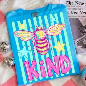Puede incluir: Una camiseta azul con un dise&ntilde;o de abeja grande, rayas verticales y estrellas. La palabra "KIND" est&aacute; impresa en rosa. La camiseta es de material Comfort Colors.