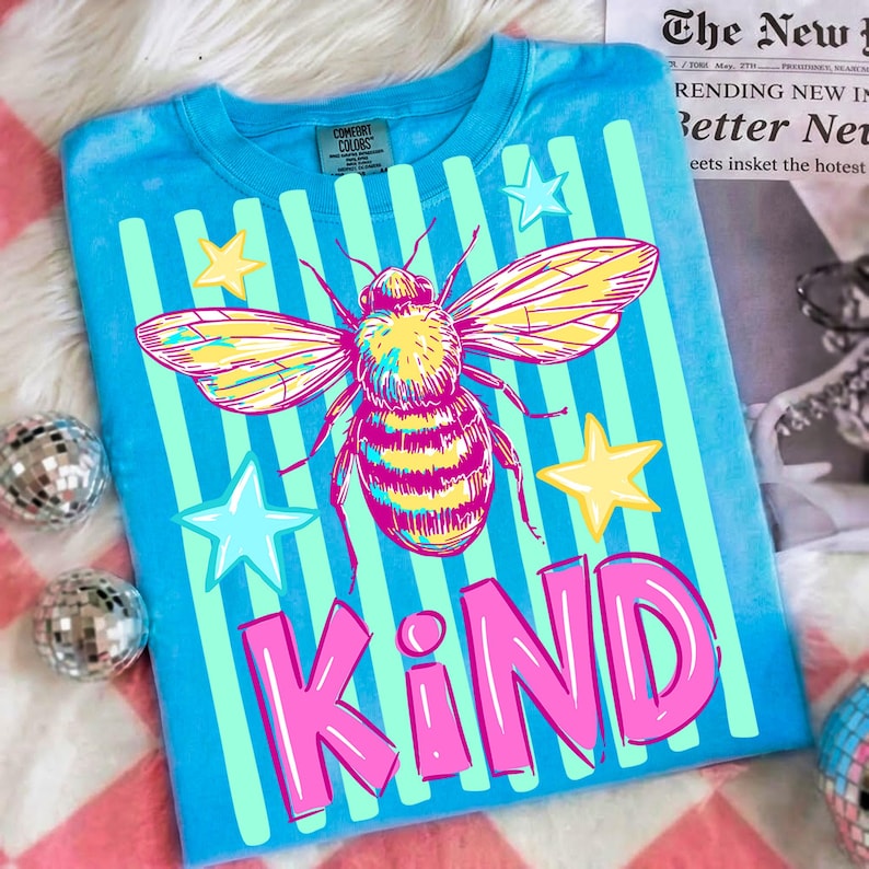 Puede incluir: Una camiseta azul con un dise&ntilde;o de abeja grande, rayas verticales y estrellas. La palabra "KIND" est&aacute; impresa en rosa. La camiseta es de material Comfort Colors.