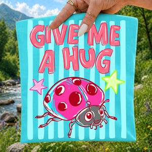 Op de afbeelding: Een turquoise draagtas met verticale lichtblauwe strepen en de tekst "GIVE ME A HUG" in roze. Een roze lieveheersbeestje met rode stippen en een groene ster staan ook op de tas.